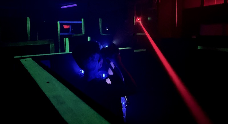 Obrázek ze hry laser game. Hráč skrčený za překážkou a nad ním laser jiného hráče, který po něm střílel.